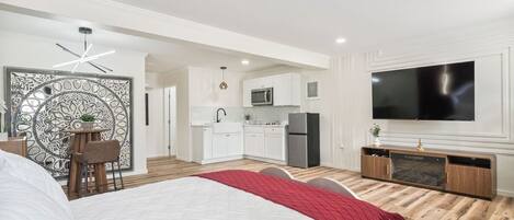 A modern bedroom with a large bed, a dining area, a kitchenette, and a TV mounted on the wall.