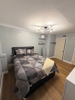 A bedroom with a bed, a bedside table, a nightstand, a wall-mounted shelf with decorative items, a door, and a ceiling light fixture. - Stylish 2-bedroom apartment with AC in near FLL airport & Hollywood beach (Hollywood)