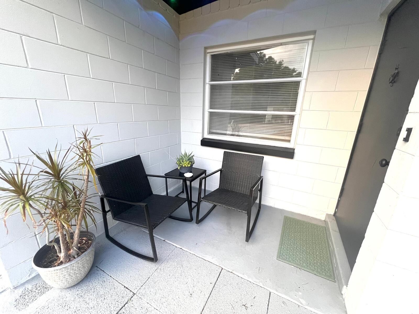 Un petit espace extérieur aménagé avec deux chaises et une table, un pot de fleurs et une fenêtre avec des stores.