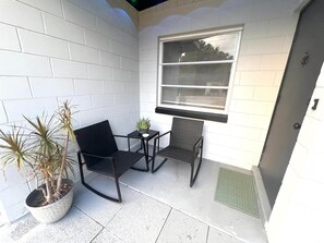 A small outdoor seating area with two chairs and a table, a potted plant, and a window with blinds.
