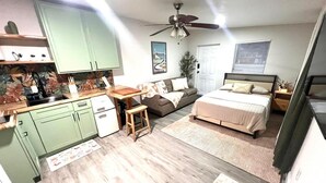 Un espacio habitable compacto con kitchenette, cama, ventilador de techo y ventana con persianas.