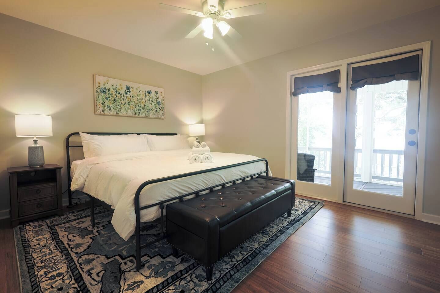 Quarto com cama, mesas de cabeceira, um banquinho, ventilador de teto, pintura e porta de vidro deslizante para área externa.