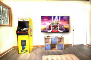 Une pièce avec une borne d’arcade Pac-Man, un téléviseur fixé au mur et une petite étagère en bois sur laquelle repose une manette de jeu.