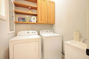 Une buanderie équipée de deux lave-linge Whirlpool blancs, d’une étagère en bois au-dessus pour ranger les produits de nettoyage, et d’un petit plan de travail avec un distributeur de savon.