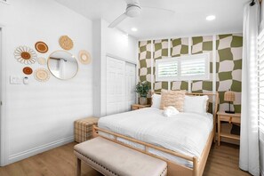 A bedroom with a bed, a bench, a ceiling fan, a wall-mounted mirror, and a window with blinds. - Fun & Modern Summer Pool Home near Disney! (Anaheim)