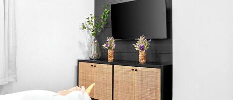 Una habitación moderna con televisión de pantalla plana montada en una pared oscura, una mesita consola de mimbre con dos jarrones y un peluche de unicornio sobre la cama.