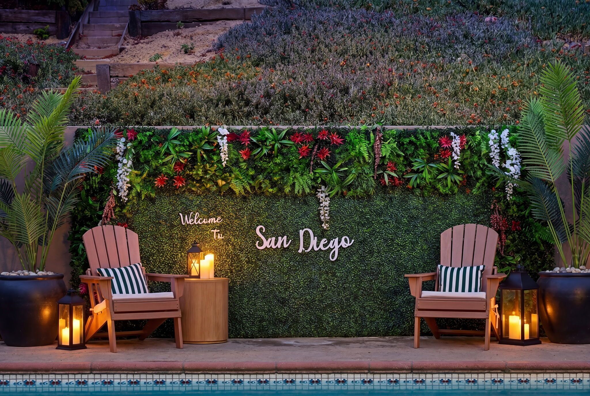 A poolside area with two wooden chairs, a small table with a candle, and a wall decorated with greenery and flowers.