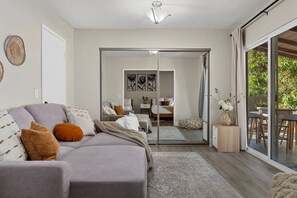 A modern living room with a grey sofa, a dining area with a table and chairs, and a sliding glass door leading to a bedroom with a bed and a small table. - San Diego Pool Home Oasis with Backyard Retreat (San Diego)