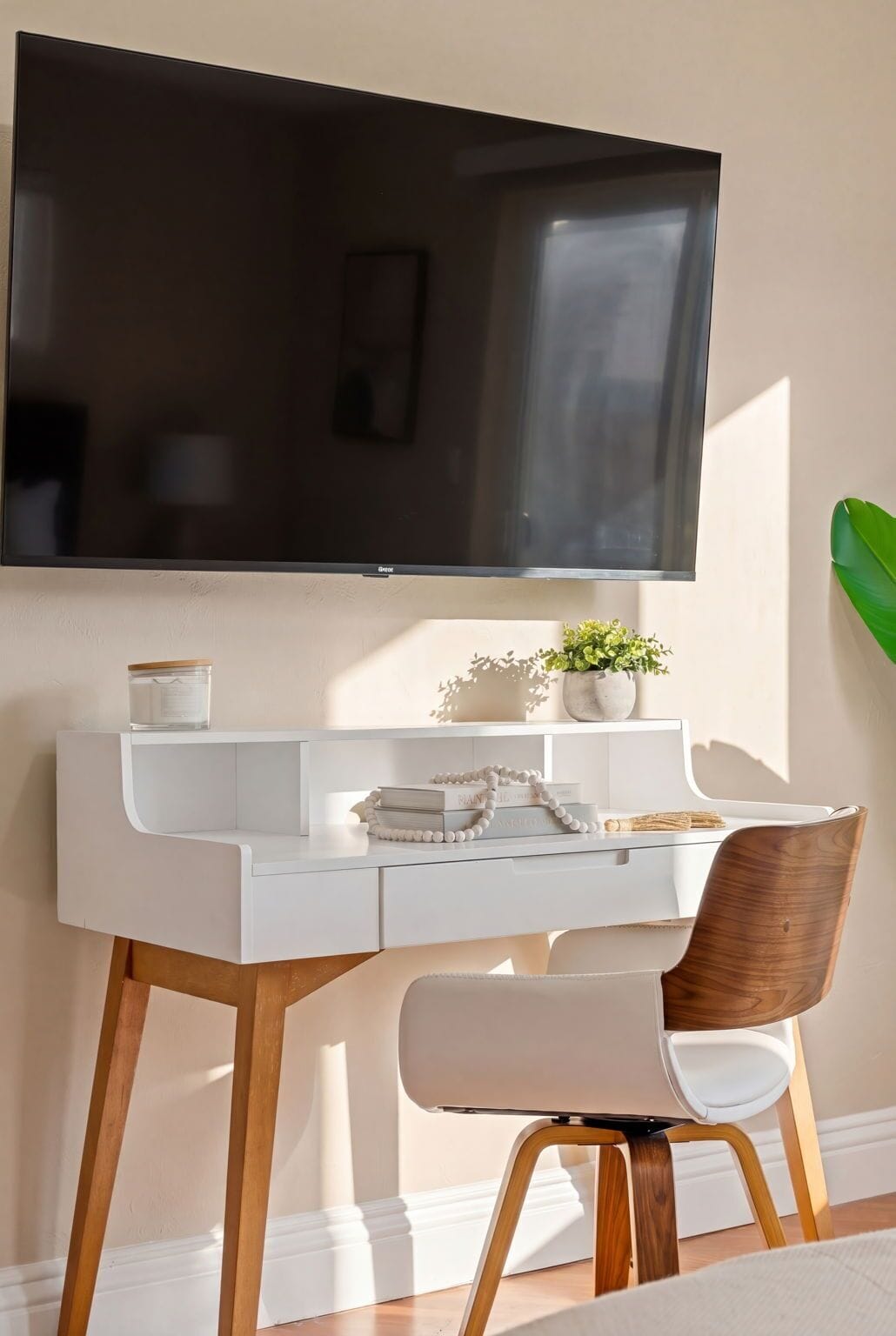 A modern desk with a flat-screen TV mounted above it, a white chair with wooden legs, and a small potted plant on the desk.
