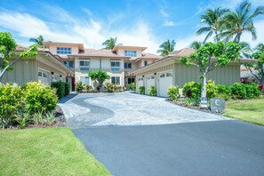 A residential building with a driveway leading to a gated entrance, surrounded by well-maintained landscaping and palm trees. - Walk to the Beach, Shopping, and Dining. (Waikoloa Village)
