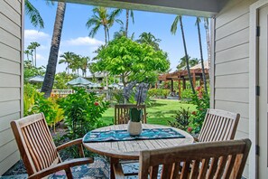 A covered patio with a wooden table and chairs, surrounded by lush greenery and palm trees. - Walk to the Beach, Shopping, and Dining. (Waikoloa Village)