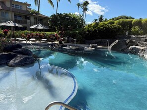 A clear pool with a blue edge, surrounded by rocks and palm trees, with a building in the background. - Walk to the Beach, Shopping, and Dining. (Waikoloa Village)