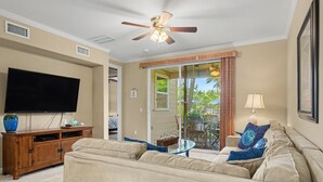 A living room with a flat-screen TV, a ceiling fan, a sofa, a coffee table, and a sliding glass door leading to an outdoor area. - Walk to the Beach, Shopping, and Dining. (Waikoloa Village)