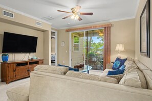 Ein Wohnzimmer mit Flachbildfernseher, Deckenventilator, Sofa, Couchtisch und einer Glasschiebetür, die zu einem Außenbereich führt.
