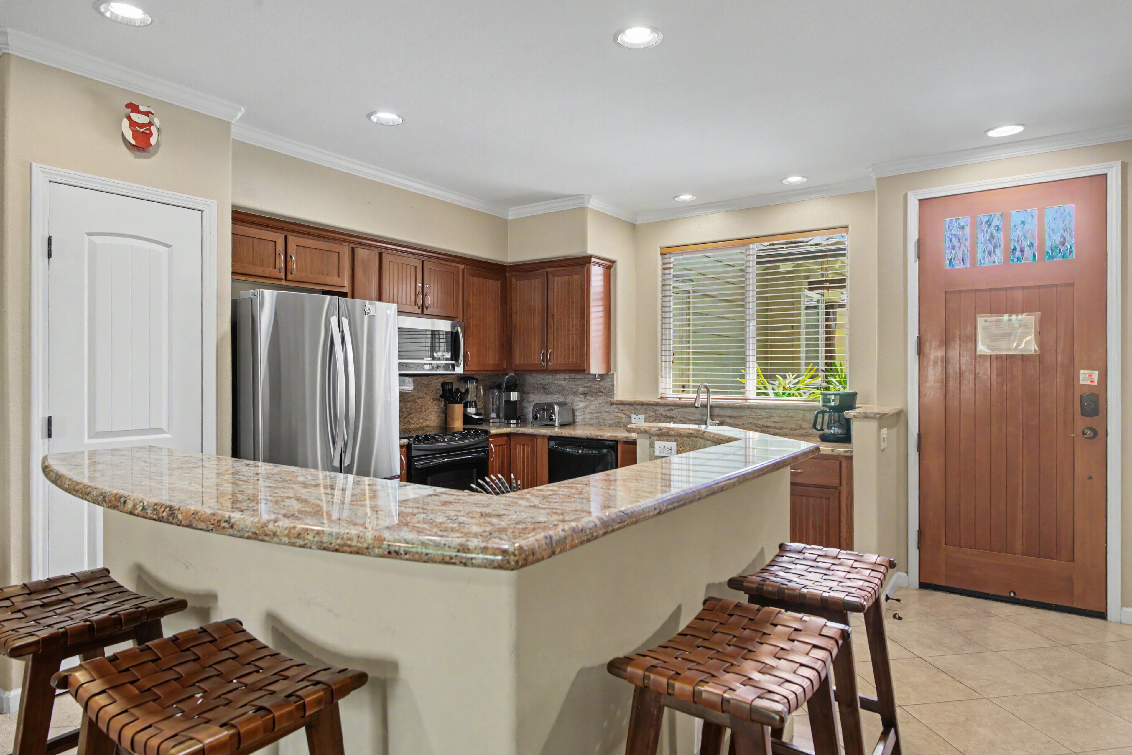 A kitchen with granite countertops, wooden cabinets, a stainless steel refrigerator, a stove, and a sink. There are four stools at the island.