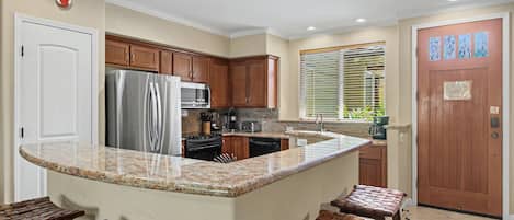 A kitchen with granite countertops, wooden cabinets, a stainless steel refrigerator, a stove, and a sink. There are four stools at the island.