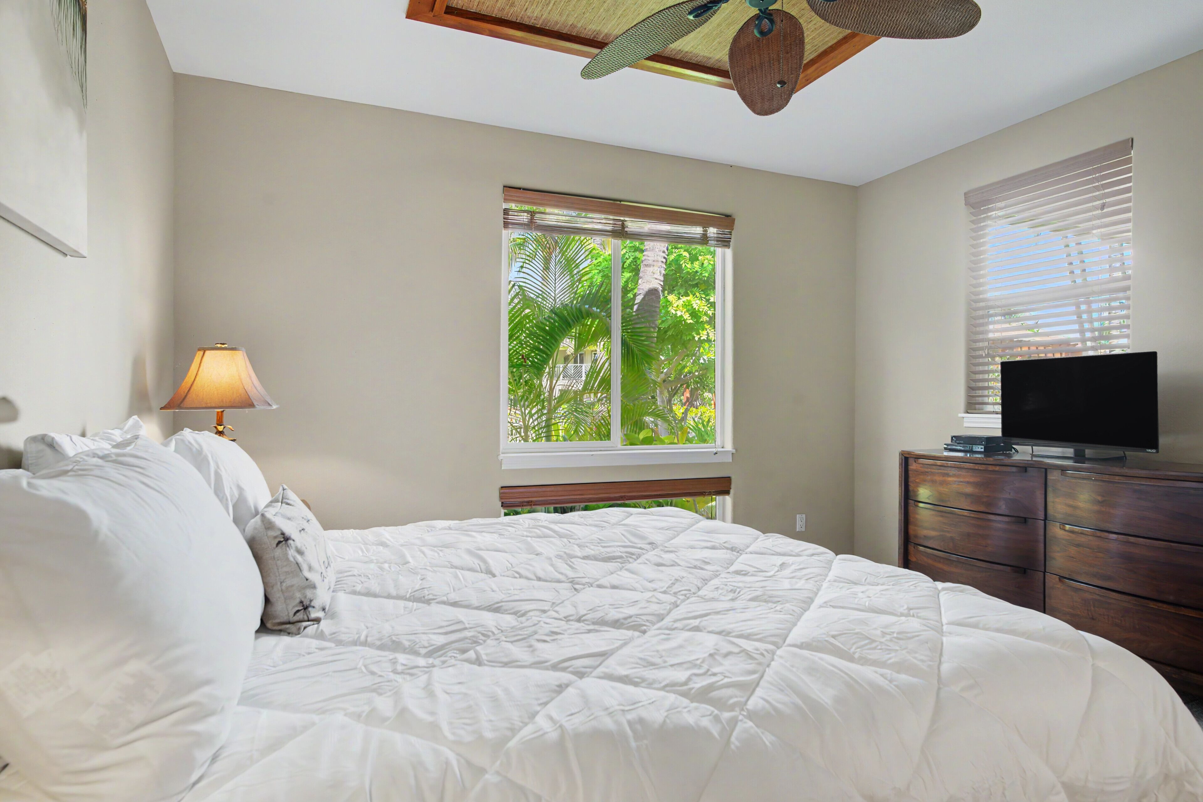 Quarto com cama, abajur, televisão sobre uma cômoda de madeira e janela com persianas.
