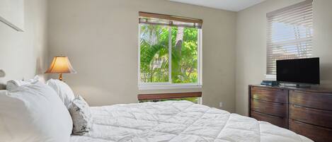 Quarto com cama, abajur, televisão sobre uma cÓmoda de madeira e janela com persianas.