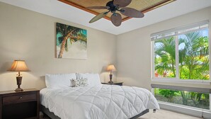 A bedroom with a bed, two nightstands, a ceiling fan, a window with a view of palm trees, and a painting of a palm tree. - Walk to the Beach, Shopping, and Dining. (Waikoloa Village)