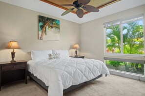 A bedroom with a bed, two nightstands, a ceiling fan, a window with a view of palm trees, and a painting of a palm tree.