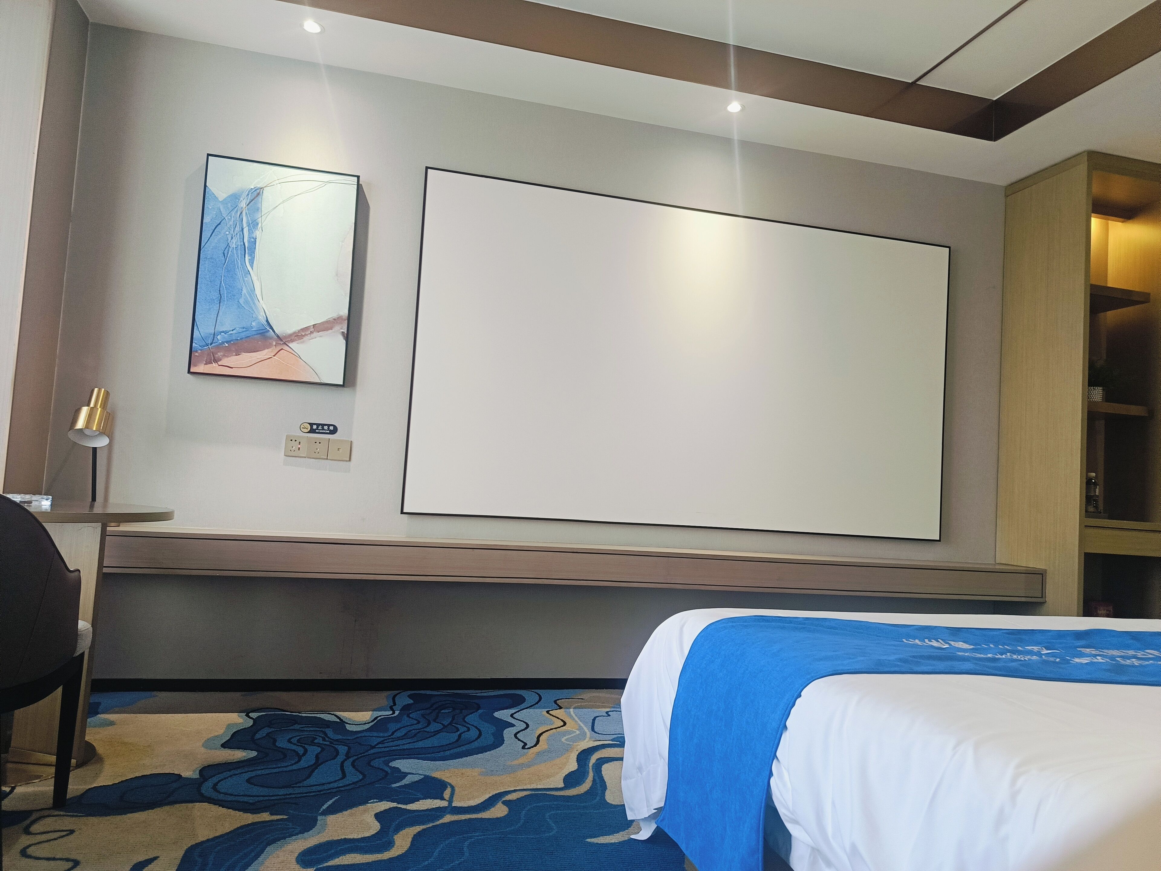 A hotel room with a bed, a desk, a chair, a lamp, and artwork on the wall.