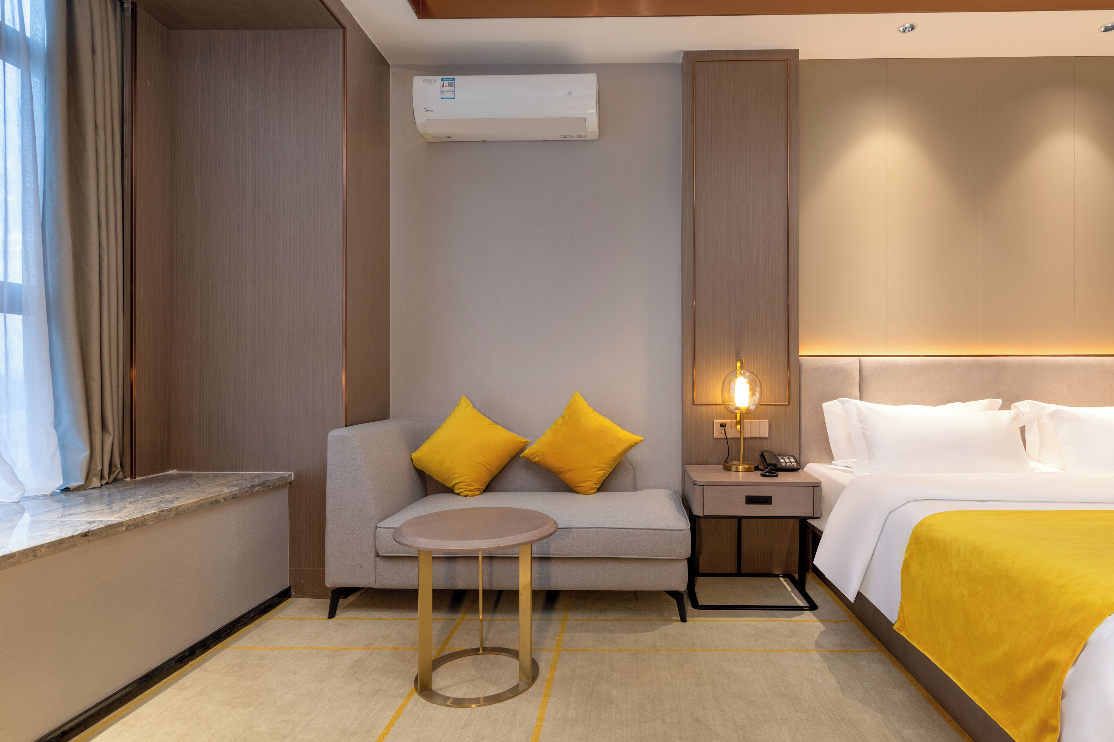 Habitación de hotel con cama, sofá con cojines amarillos, una mesita redonda, mesita de noche con lámpara y un equipo de aire acondicionado en la pared.