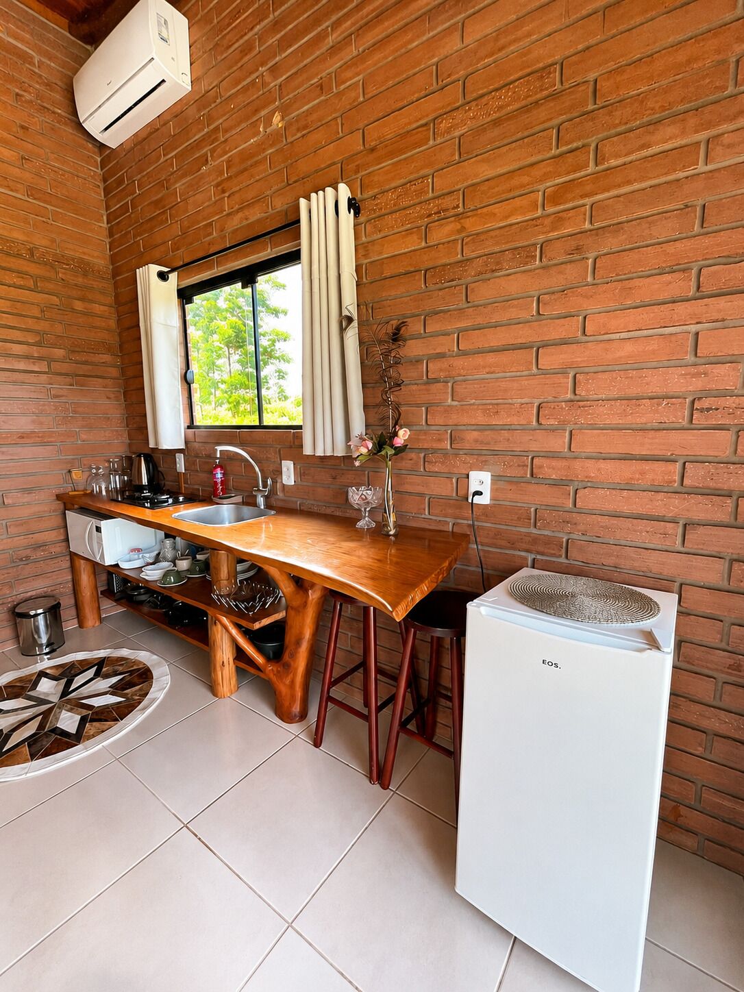 A compact kitchen with a brick wall, a wooden table, a white refrigerator, and a window with curtains.