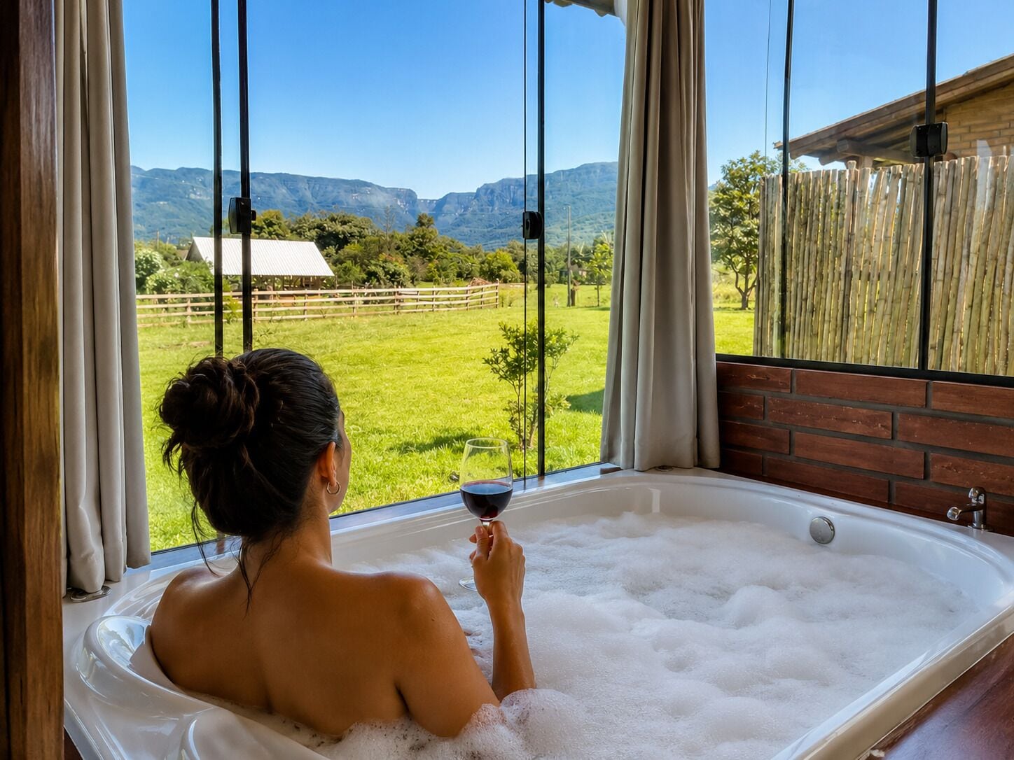 Una persona relajándose en una bañera con una copa de vino tinto, con vistas a un paisaje verde y montañas al fondo.
