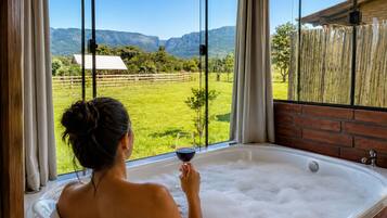 Una persona relajándose en una bañera con una copa de vino tinto, con vistas a un paisaje verde y montañas al fondo.