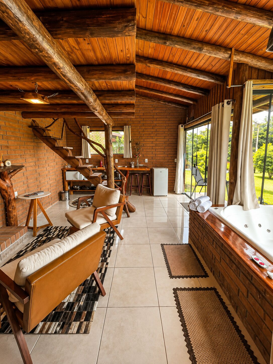 A cozy cabin interior with a wooden ceiling, brick walls, a seating area with a sofa and chairs, a dining table, and a large window offering a view of greenery.