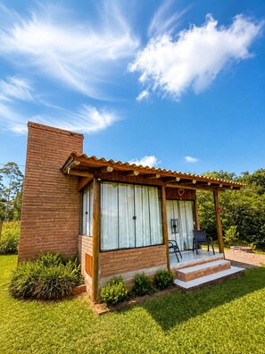 Una casa de un piso con fachada de ladrillo, un porche cubierto con dos sillas y un cielo azul despejado.