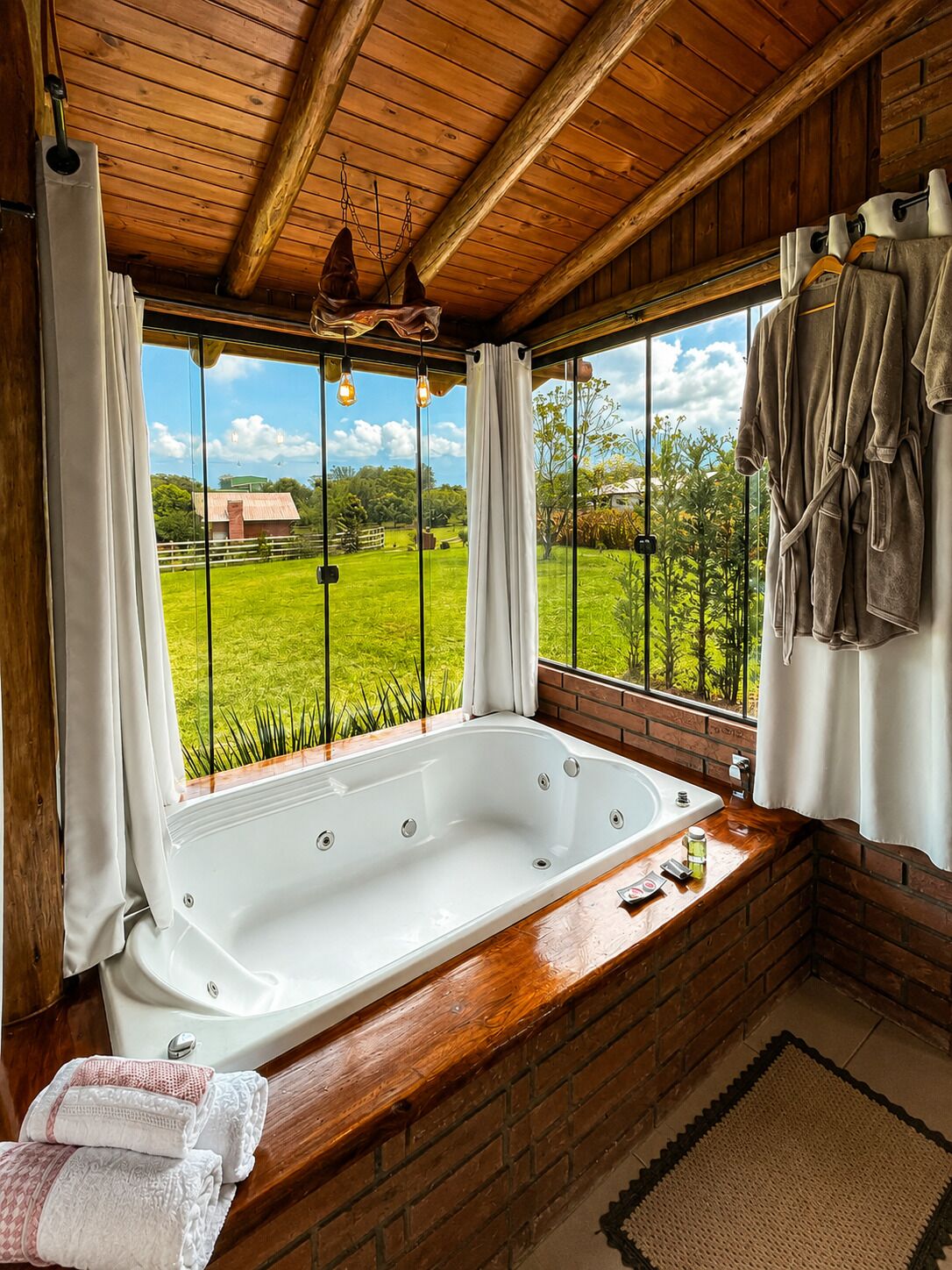 Un bagno con una grande vasca, travi in legno a soffitto e vista su un paesaggio verde attraverso ampie finestre.