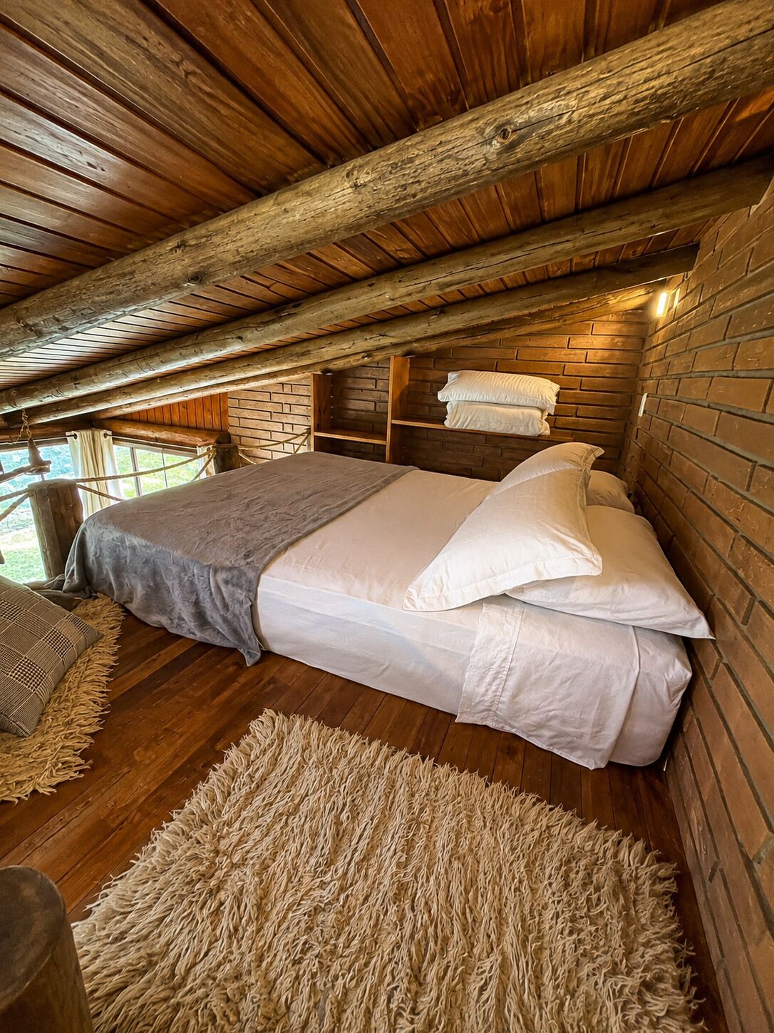 A cozy attic bedroom with a wooden ceiling, a large bed with white linens, and a rustic brick wall.