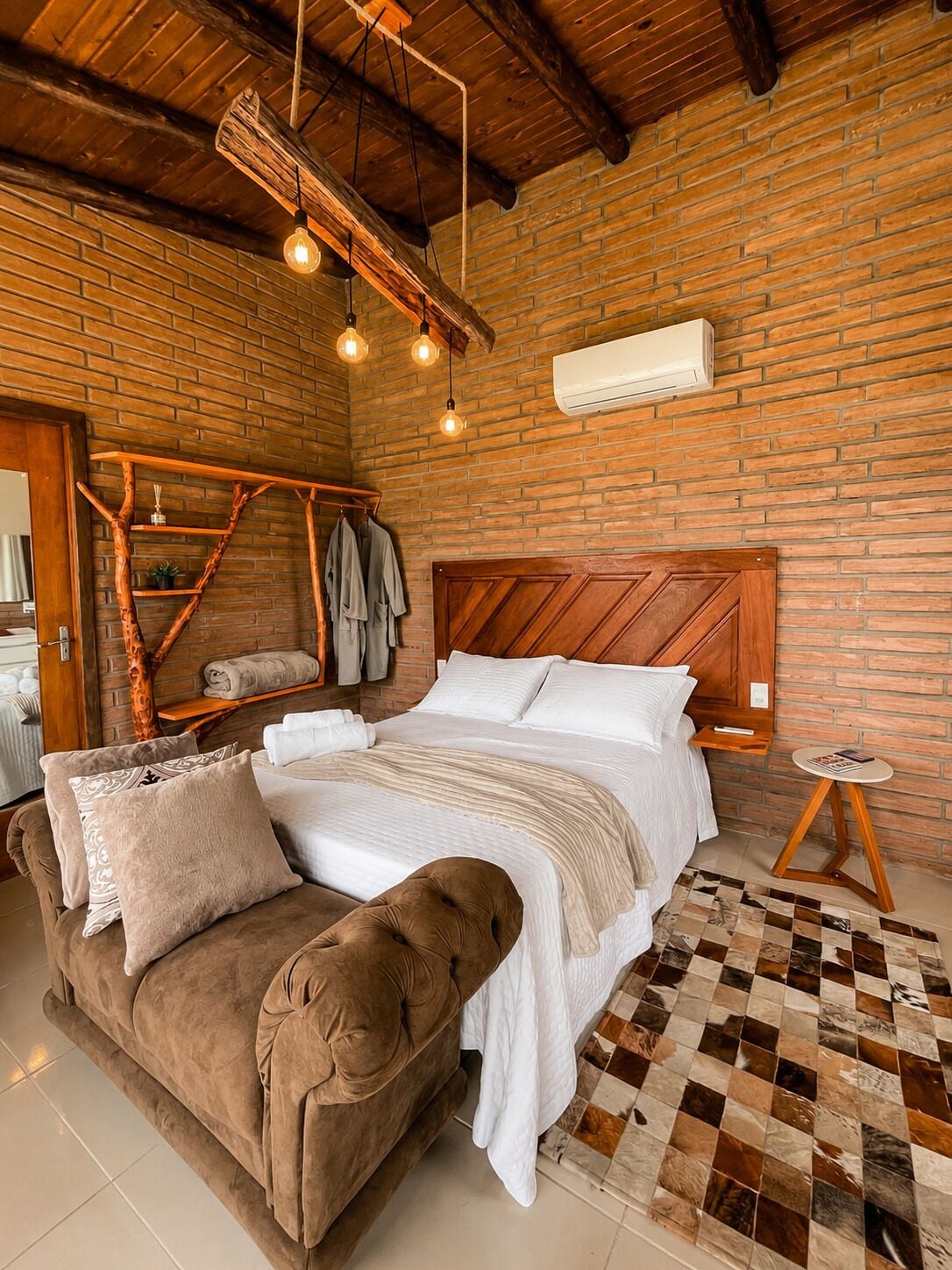 A cozy bedroom with a brick wall, a wooden headboard, a bed with white linens, a brown sofa, and a small round table.