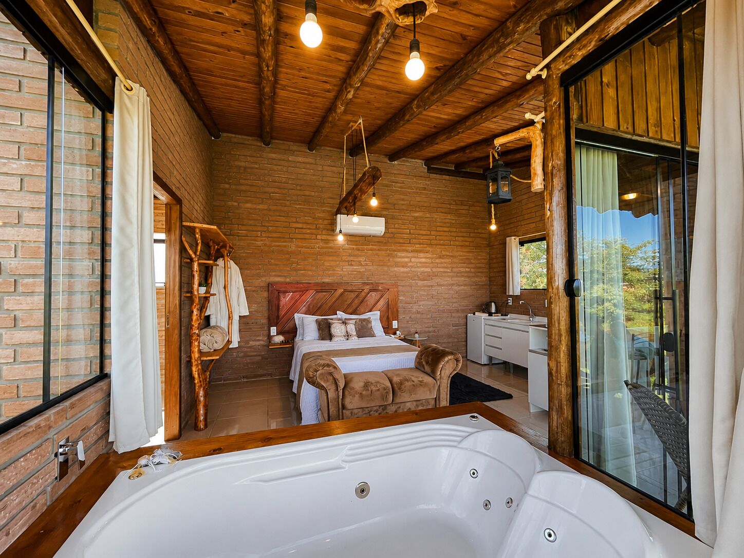 A room with a large wooden ceiling, a bed with pillows, a white bathtub, and a window with curtains.