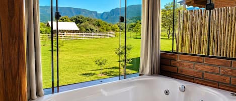 A bathroom with a large window, a bathtub, and a view of a green field and mountains.