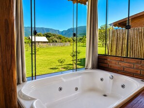 A bathroom with a large window, a bathtub, and a view of a green field and mountains.
