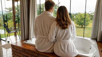 Ein Paar in weißen Bademänteln sitzt auf einer hölzernen Bank vor einem großen Fenster mit Blick auf eine Landschaft.