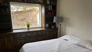 A bedroom with a bed, a window with a view of a stone wall, a bookshelf, and a lamp.