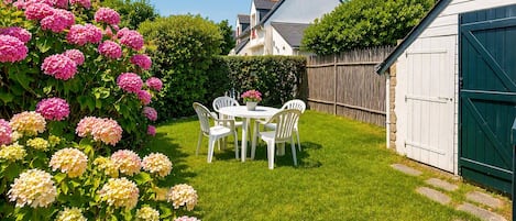 A well-maintained garden with a white table and chairs, surrounded by lush greenery and flowering plants.