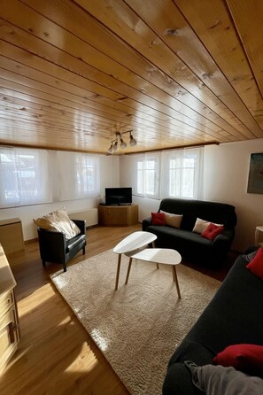 Een gezellige woonkamer met houten balken, een zwarte bank, een witte salontafel en een televisie.