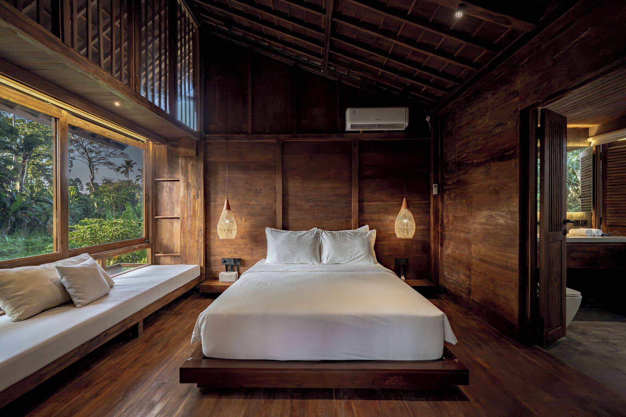 A large bed with white linens in a wooden room with a window and two hanging lamps.