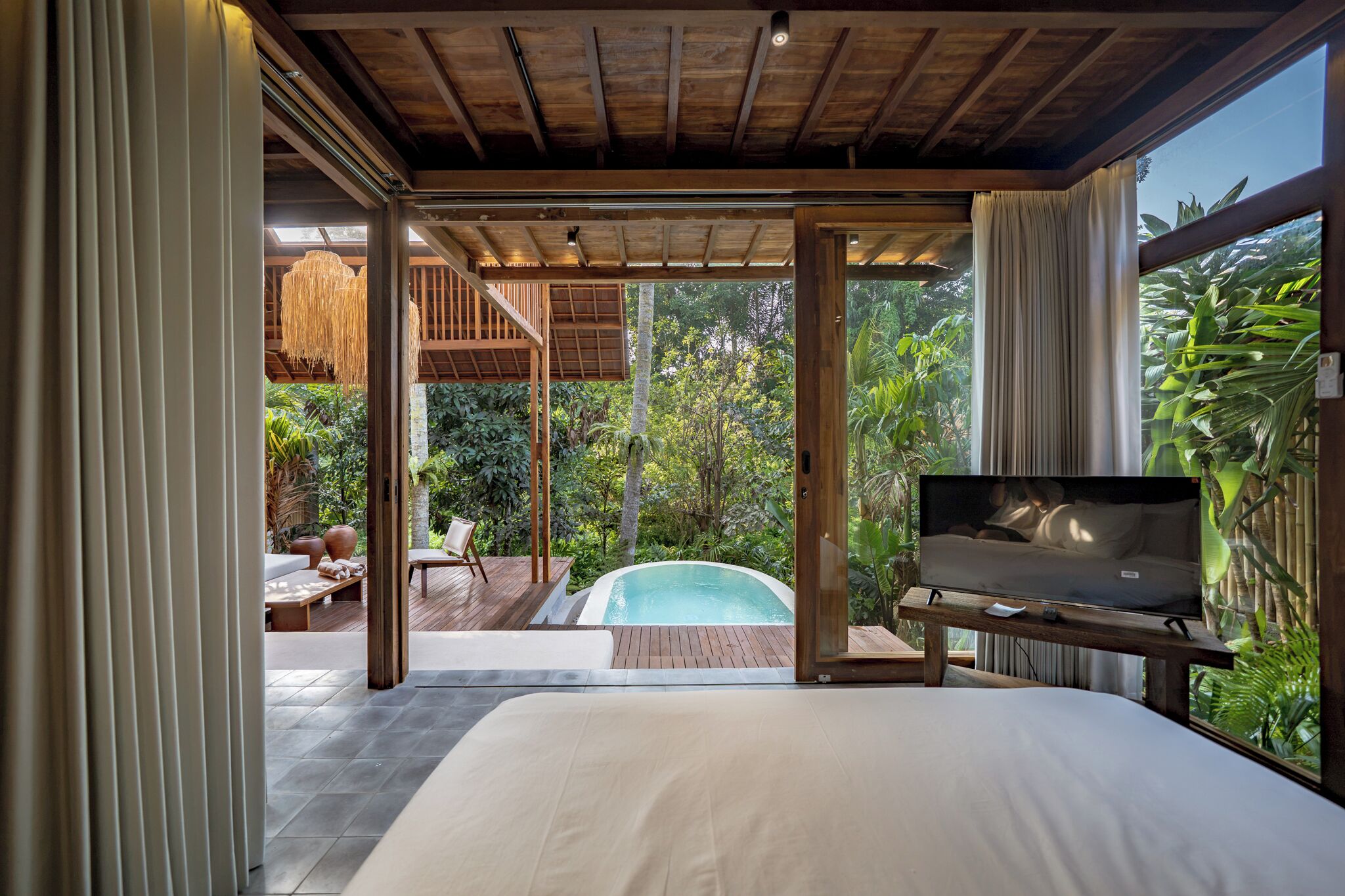 A bedroom with a large bed, a TV, and a view of a swimming pool and lush greenery.