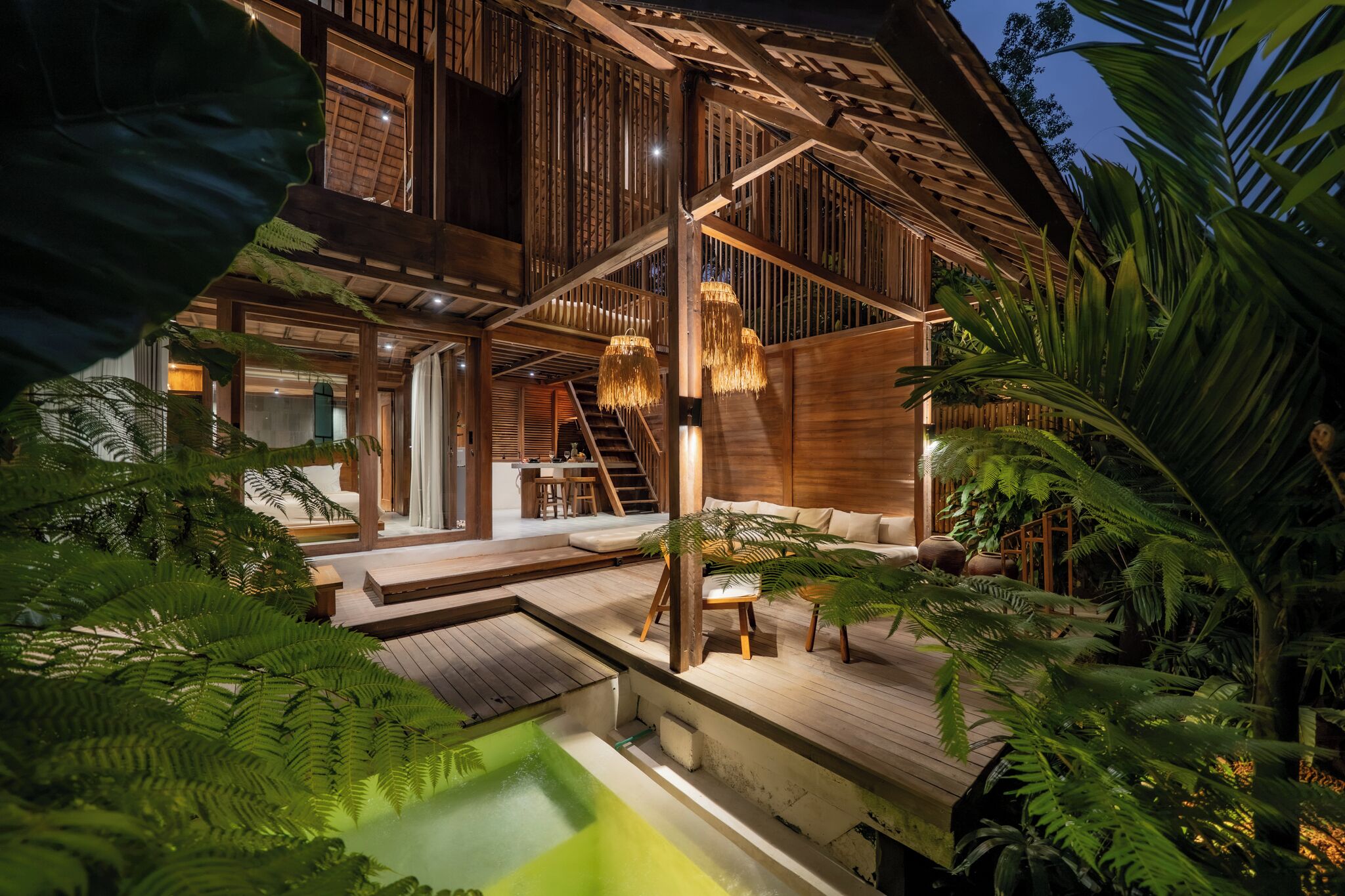 A wooden house with a pool, staircase, and outdoor seating area surrounded by lush greenery.
