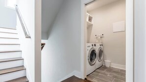 Ein Waschraum mit Waschmaschine und Trockner, einem kleinen Mülleimer und einer weißen Wand mit Lichtschalter.