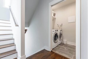 A laundry room with a washing machine and dryer, a small trash bin, and a white wall with a light switch. - 4BR East Nashville - Walk to Bars & Restaurants (Nashville)