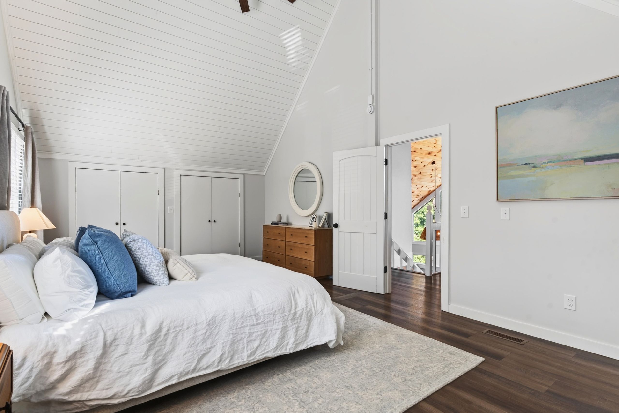 Ein Schlafzimmer mit schrägem Dach, einem Bett mit weißer Bettwäsche, einer Holzkommode, einem runden Spiegel und einem Bild an der Wand.
