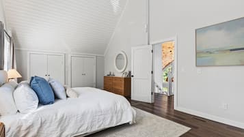 Ein Schlafzimmer mit schrägem Dach, einem Bett mit weißer Bettwäsche, einer Holzkommode, einem runden Spiegel und einem Bild an der Wand.