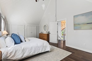 Ein Schlafzimmer mit schrägem Dach, einem Bett mit weißer Bettwäsche, einer Holzkommode, einem runden Spiegel und einem Bild an der Wand.