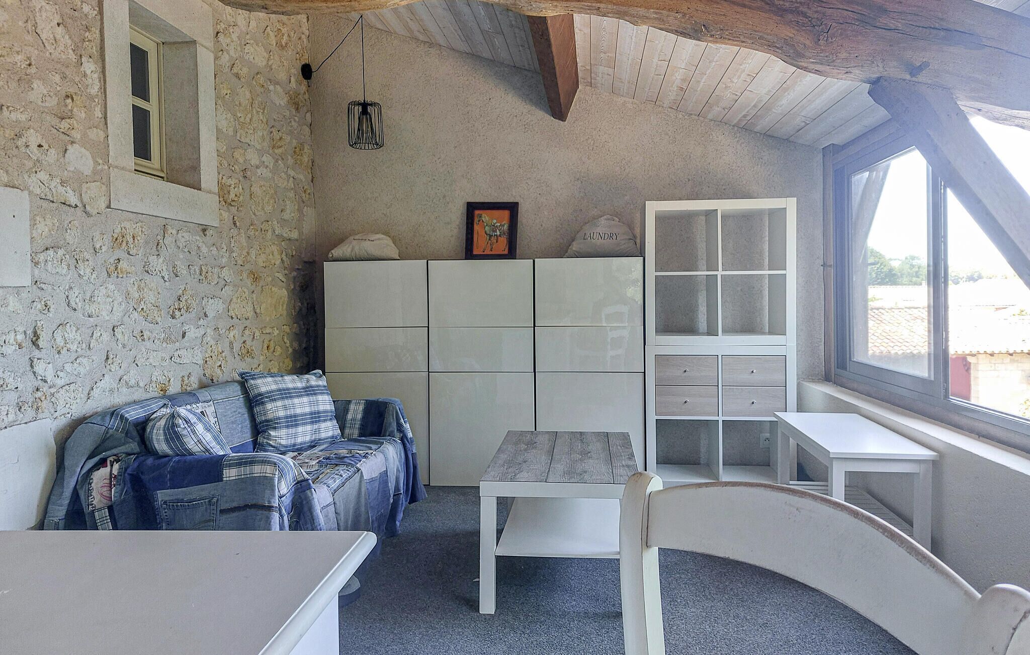 Une pièce avec un mur en pierre, un plafond en bois, un canapé recouvert d’une couverture à carreaux, une commode blanche avec des tiroirs et une fenêtre donnant sur un beau paysage.